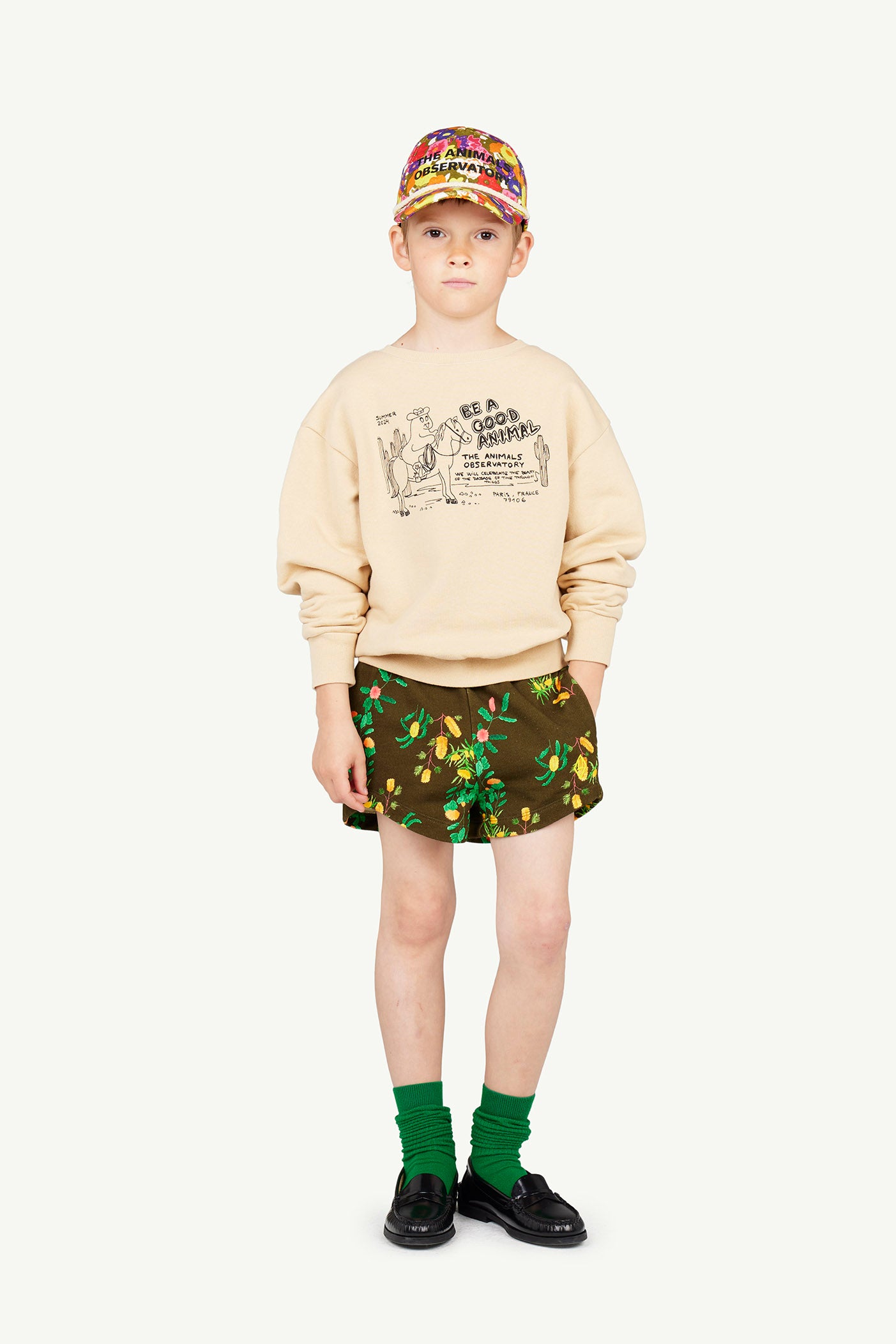 Image of a boy dressed in The Animals Observatory. The outfit consists of a cap, a sweatshirt, printed shorts, and green socks. The image shows the outfit from the front.